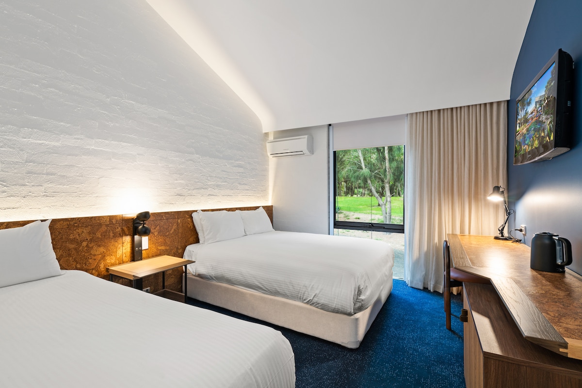 A spacious guest room features a queen bed and a single bed, with soft white linens and wooden accents. Natural light enters through a large window, providing a view of greenery outside. A desk and chair are positioned thoughtfully near the window.