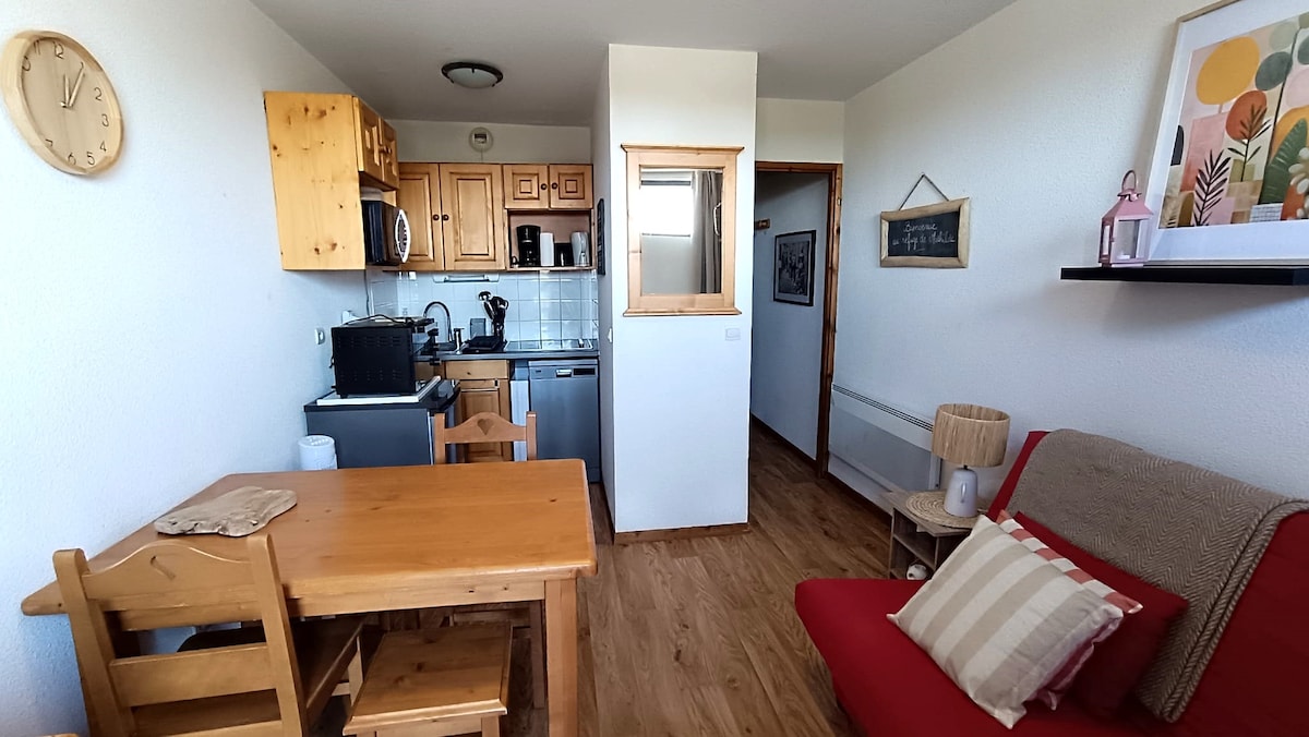 A cozy living area features a wooden table surrounded by chairs, a red sofa with decorative cushions, and a kitchenette with essential appliances. Natural light enters from a nearby window, illuminating the space. Simple decor accentuates the warm tones of the wooden cabinetry.