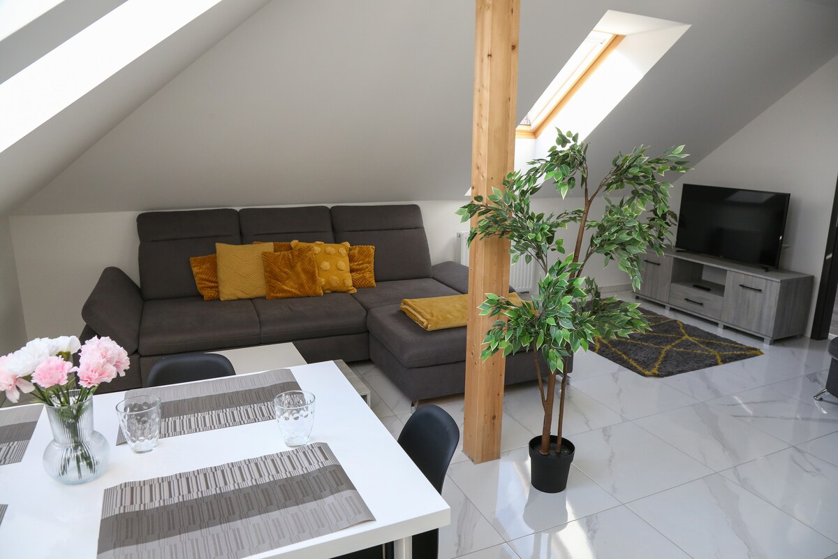 A bright living space features a sectional sofa adorned with mustard-colored cushions. A small dining area showcases a white table set with glassware, while a potted plant adds greenery. Natural light streams through skylights, enhancing the modern design.