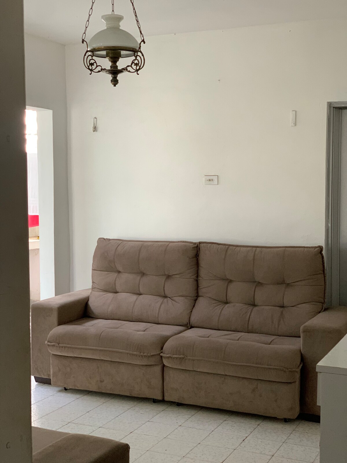A neutral-colored sofa with a tufted design is positioned against a white wall. A vintage-style light fixture hangs from the ceiling, providing soft illumination. The setting is minimalistic, contributing to a spacious feel within the room.