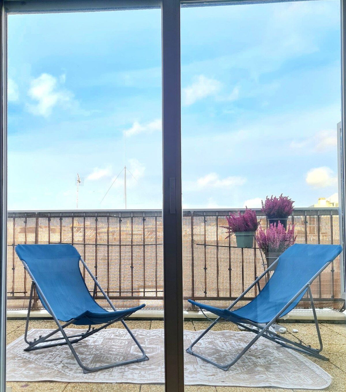 A private terrace is depicted, featuring two blue folding chairs positioned on a patterned rug. Potted plants with purple flowers are arranged near the railing, while a clear sky with soft clouds is visible in the background.