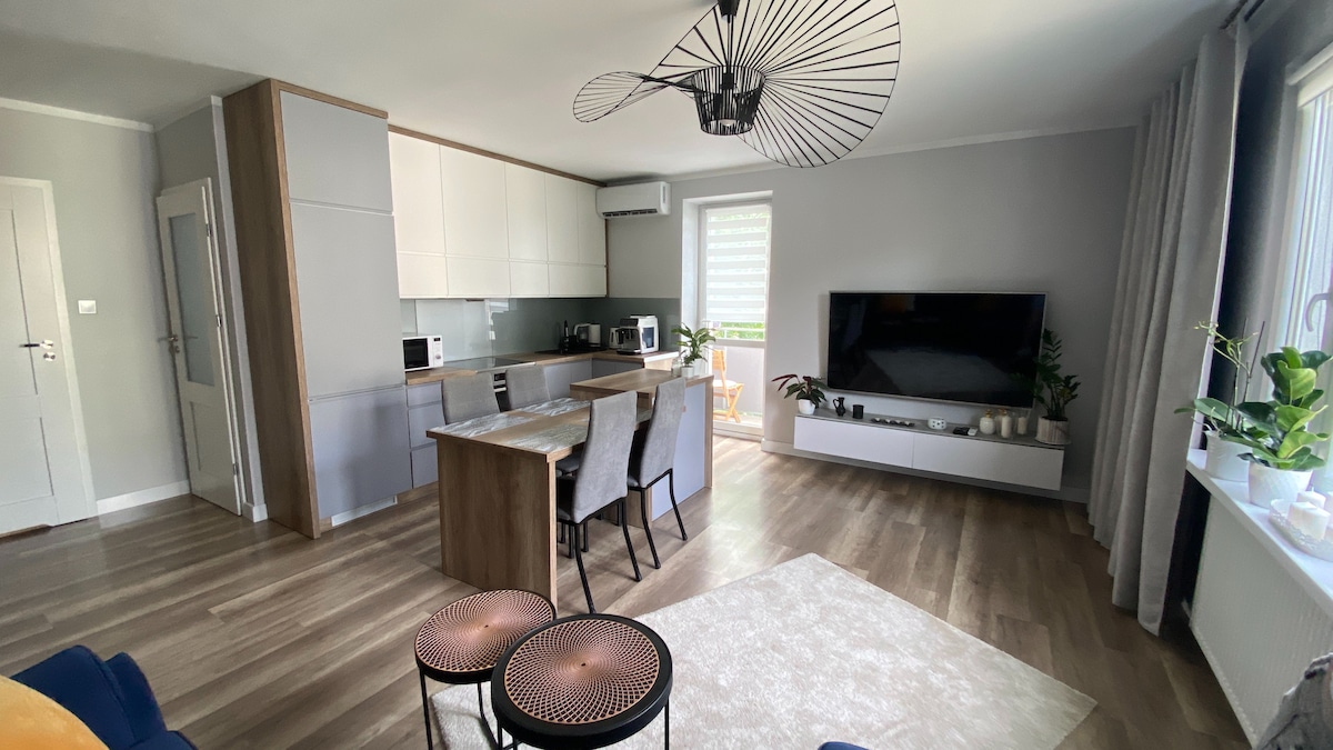 A modern living space is presented, featuring a kitchen area equipped with a refrigerator and microwave. A dining table with four chairs is positioned near a large window, with a comfortable seating area and a television mounted on the wall. Light wooden floors enhance the ambiance.