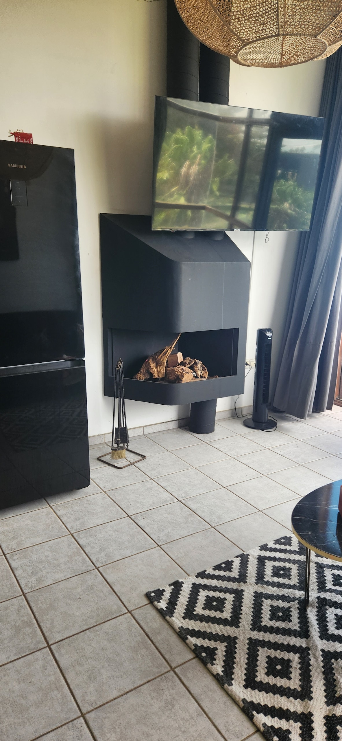 A modern fireplace is prominently featured, with logs arranged inside. Above it, a flat-screen television is mounted on the wall. Adjacent to the fireplace, a sleek black refrigerator stands against a tiled floor. A patterned rug adds texture to the space.