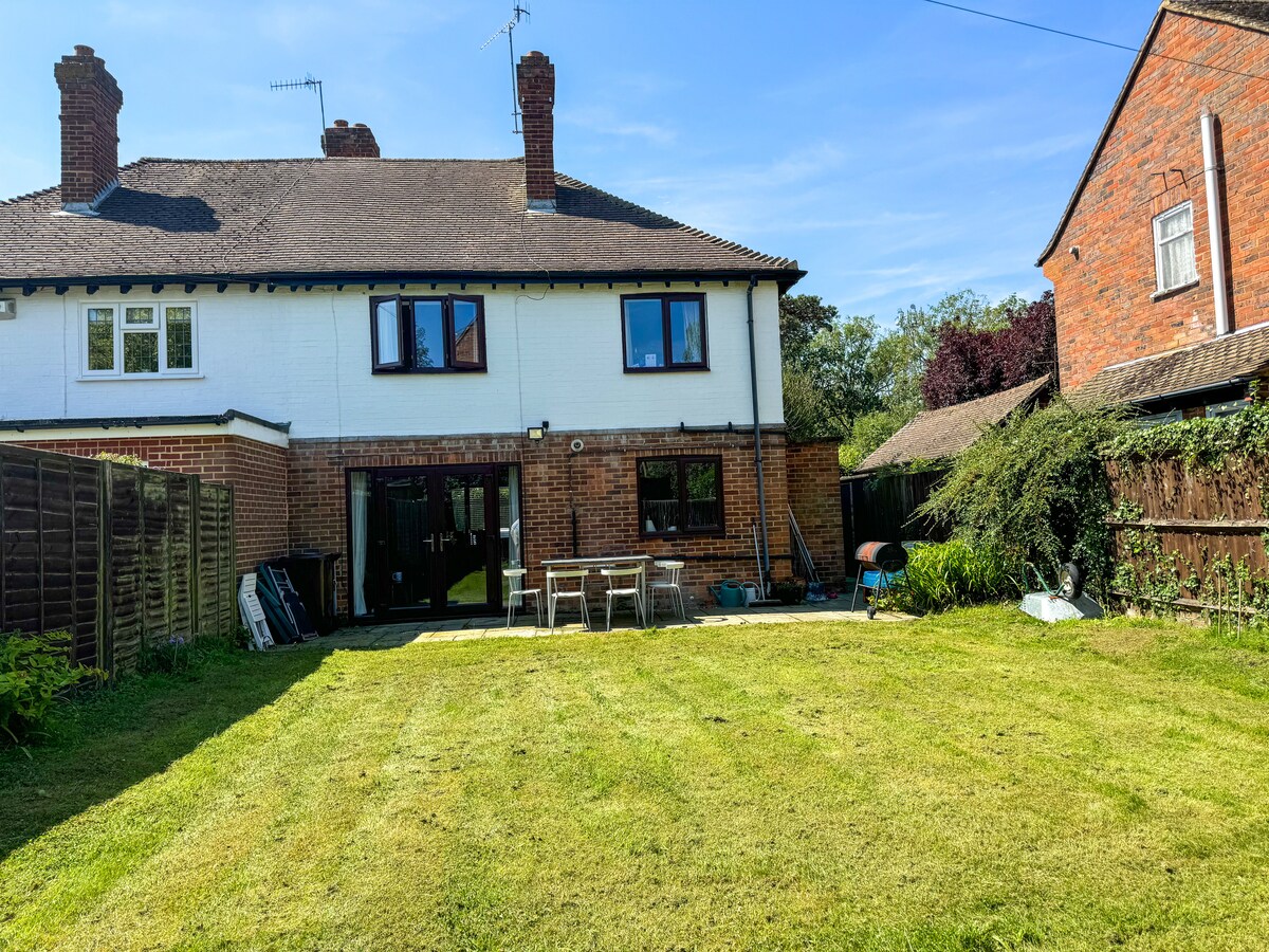 A spacious garden is presented, featuring a neatly mowed lawn and outdoor dining set positioned on a patio. A barbecue stands nearby, with various plants and trees framing the area. The back of the house showcases large glass doors, allowing for easy access to the indoor living space.