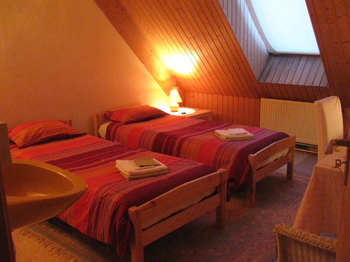 A room is presented with two beds featuring colorful striped bedspreads. Soft lighting emanates from a lamp on a nightstand. A small washbasin is visible in the corner, and a window beneath a slanted ceiling allows natural light to enter the space.