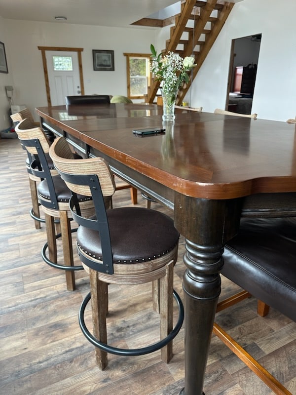 A large wooden dining table is accompanied by several high-backed chairs, featuring cushioned seats. A small glass vase with flowers decorates the center of the table. The space is brightened by natural light from nearby windows, with a staircase visible in the background.