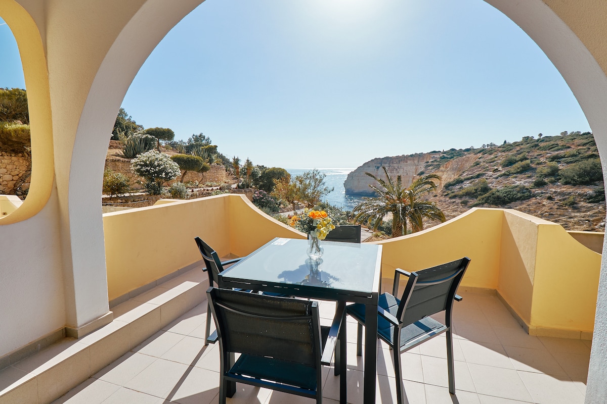 A spacious terrace overlooks the sea, featuring a dining table with four chairs. A vase with fresh flowers sits at the center of the table. The surrounding landscape includes palm trees and gently sloping hills bathed in sunlight.