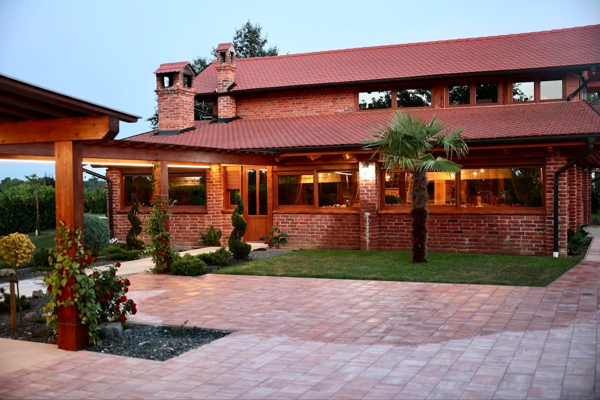 A charming brick residence with a red tile roof is surrounded by a manicured lawn. Large windows allow warm light to shine through, highlighting the garden's carefully arranged shrubs and flowers. The patio features a tiled surface, creating a welcoming outdoor area.