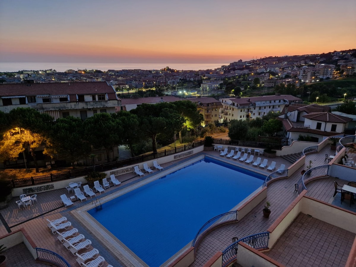 A large swimming pool is situated on a terrace, surrounded by sun loungers and trees. The evening sky is painted in soft hues as the sun sets over a hillside town, providing a serene backdrop to the tranquil scene.