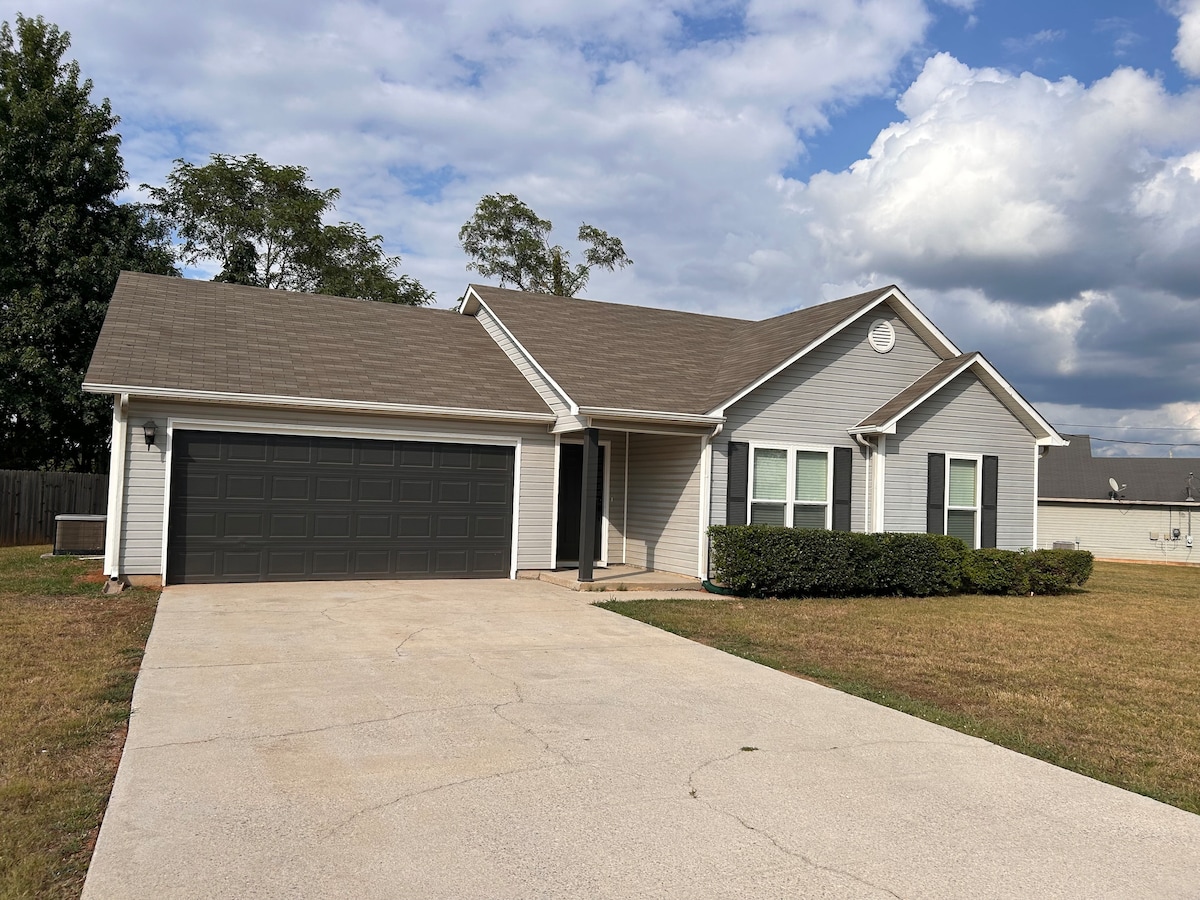 The single-story home features a neatly landscaped front yard with low-maintenance shrubbery. A spacious driveway leads to a two-car garage with a dark door. Natural light highlights the exterior, showcasing large windows and a welcoming front porch.