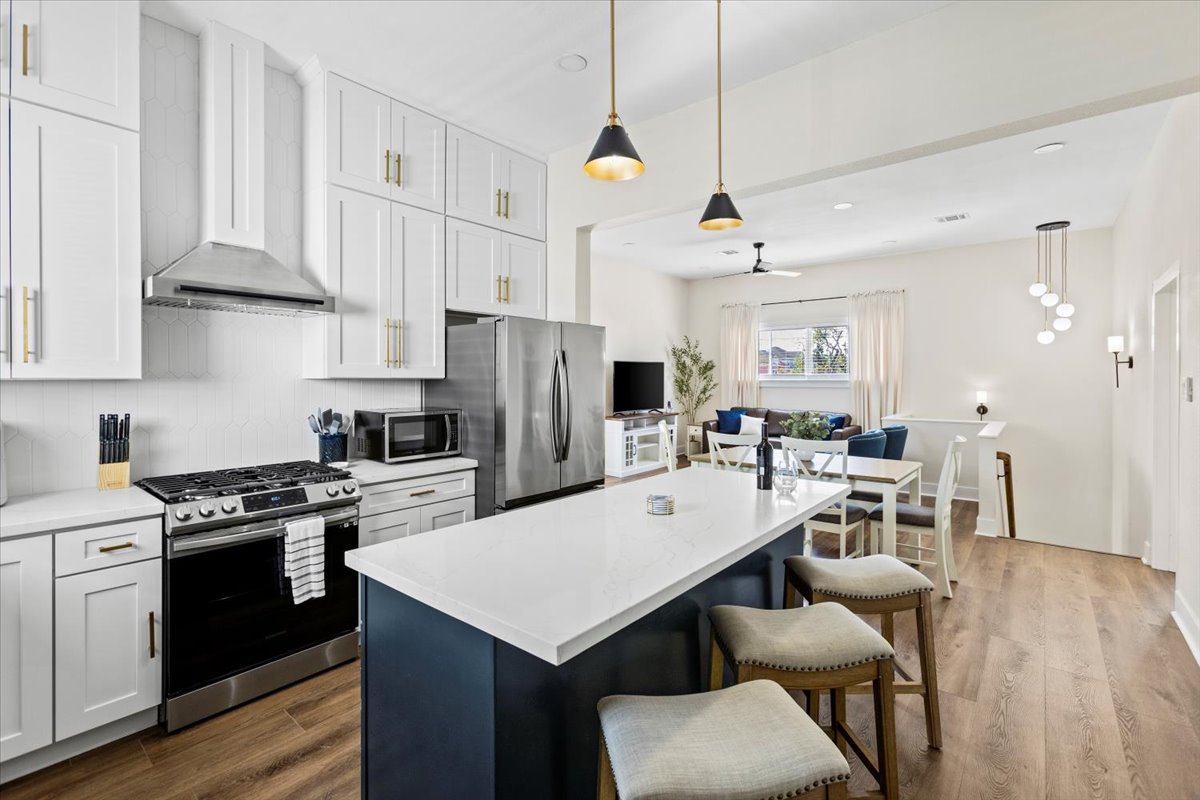 An open-concept kitchen features modern stainless steel appliances and white cabinetry. A large island with seating for three is positioned in front of a sleek glass cooktop. The adjacent living area is accented by natural light and is visible through a connecting archway.