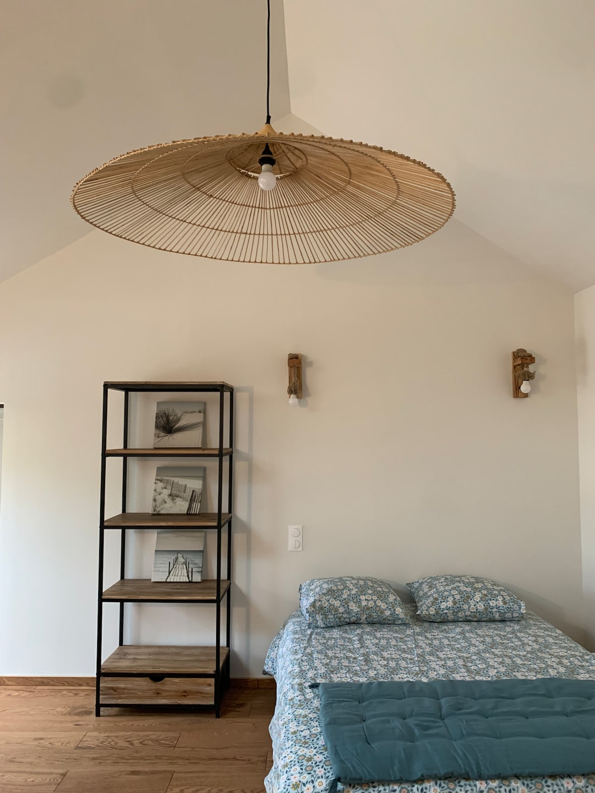 A cozy sleeping area features a spacious bed adorned with a floral patterned blanket and two soft pillows. A simple wooden shelf displays framed black-and-white photographs. A woven light fixture hangs elegantly from the ceiling, complemented by wall-mounted light sources.