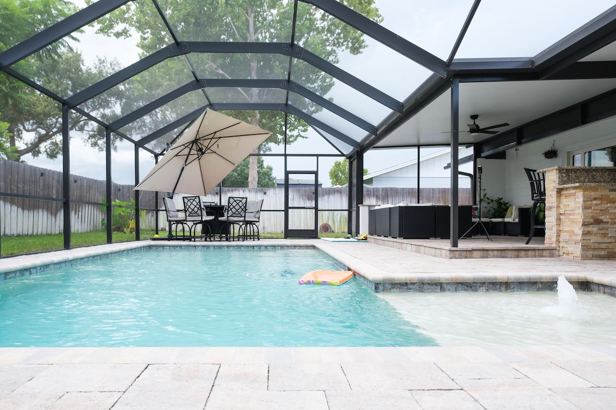 A private heated pool is enclosed by a screen and features a spacious deck. Outdoor seating is available under an umbrella, while a stone outdoor kitchen is visible nearby. Lush greenery surrounds the area, promoting a serene environment for relaxation.