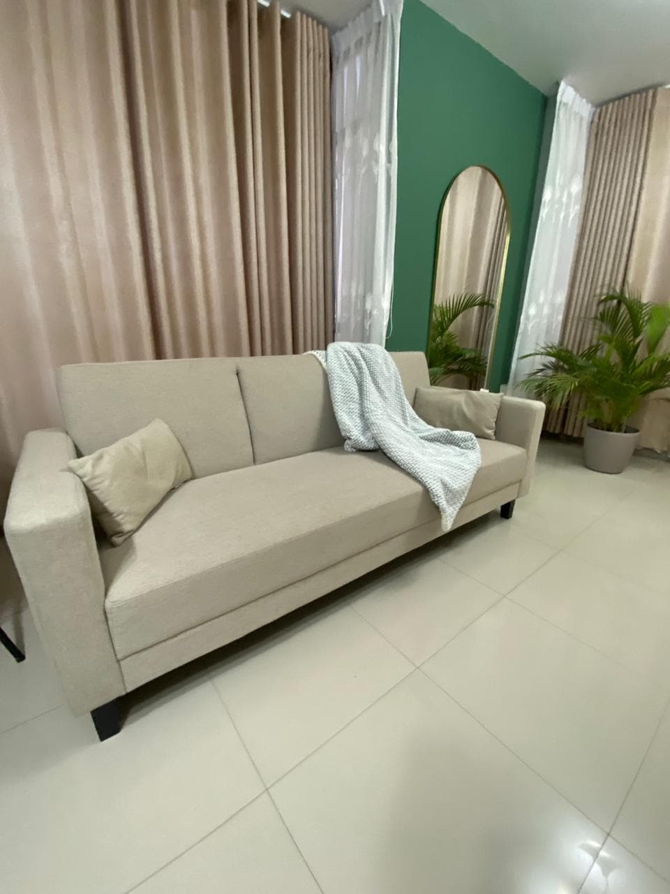 A light-colored sofa is positioned against a green accent wall, accompanied by a cozy throw draped over one arm. Soft curtains frame nearby windows, and a potted plant adds a touch of greenery to the space.