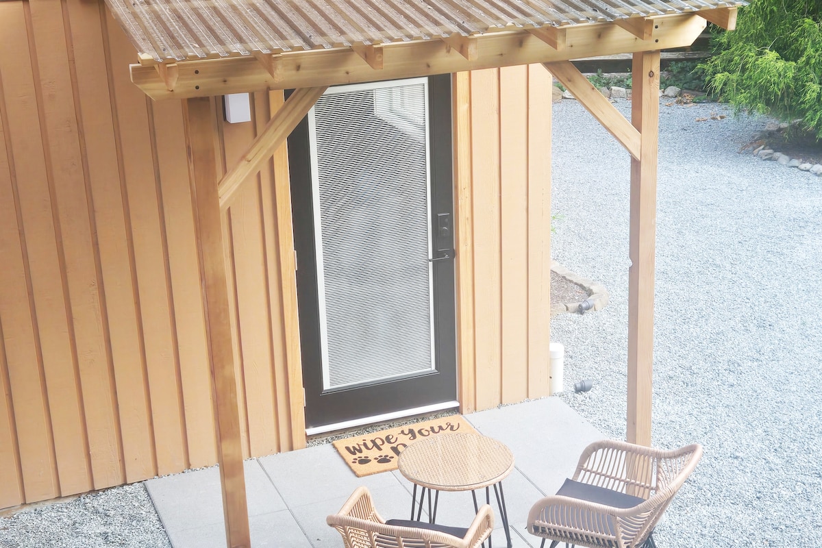 An outdoor patio area is presented, featuring a small round table and two woven chairs under a wooden awning. A decorative mat lays in front of the glass door, and gravel flooring surrounds the seating area.