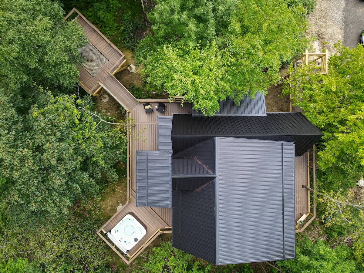 An aerial view showcases the treehouse nestled within lush greenery. Surrounding decks are visible, leading to a hot tub situated in a wooden enclosure. The design includes a combination of dark roofing and wooden accents, enhancing the integration with the natural environment.