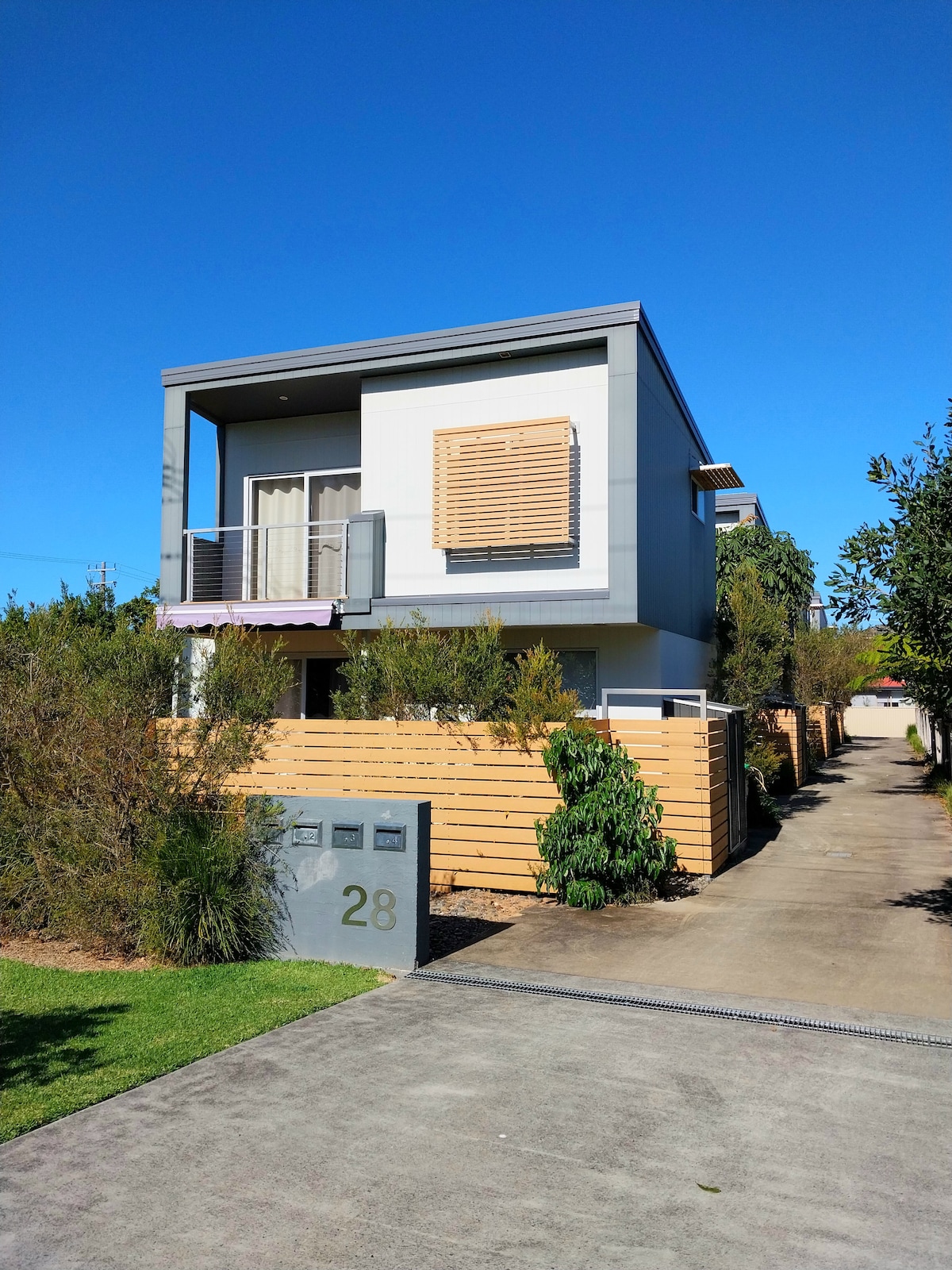 The modern townhouse is set against a clear blue sky, featuring a minimalist facade with a wooden slatted window and a covered balcony. Lush greenery and low wooden fencing provide privacy along the pathway leading to the entrance, marked by a stylish numeral.