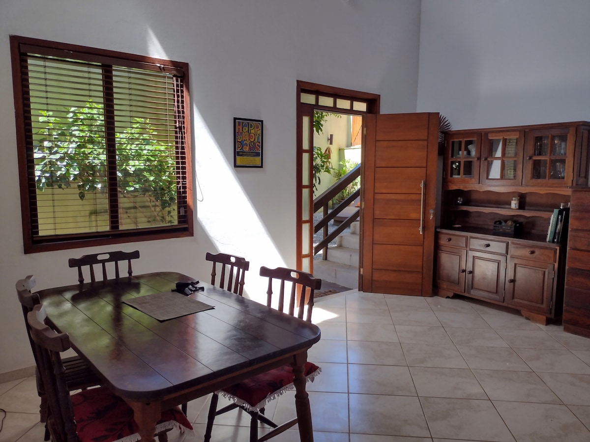 A dining area is presented with a wooden table surrounded by six chairs. Natural light filters through a window, casting shadows on the tiled floor. A wooden cabinet is positioned along one wall, showcasing a mix of textures and colors in the decor.