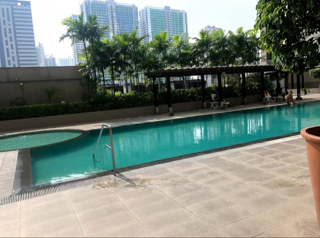 A serene outdoor pool area is presented, featuring clear blue water and a gentle staircase entry. Surrounding greenery and tall buildings in the background enhance the tranquil setting. Lounge chairs are visible nearby, providing inviting spots for relaxation.