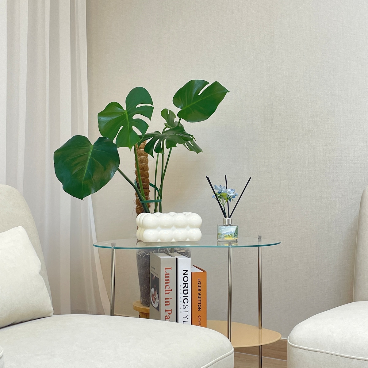 A side table is adorned with a glass top and metal legs, holding a small potted plant and a decorative item. A stack of books and a diffuser are also visible, adding a touch of decor to the calm and tidy living space.