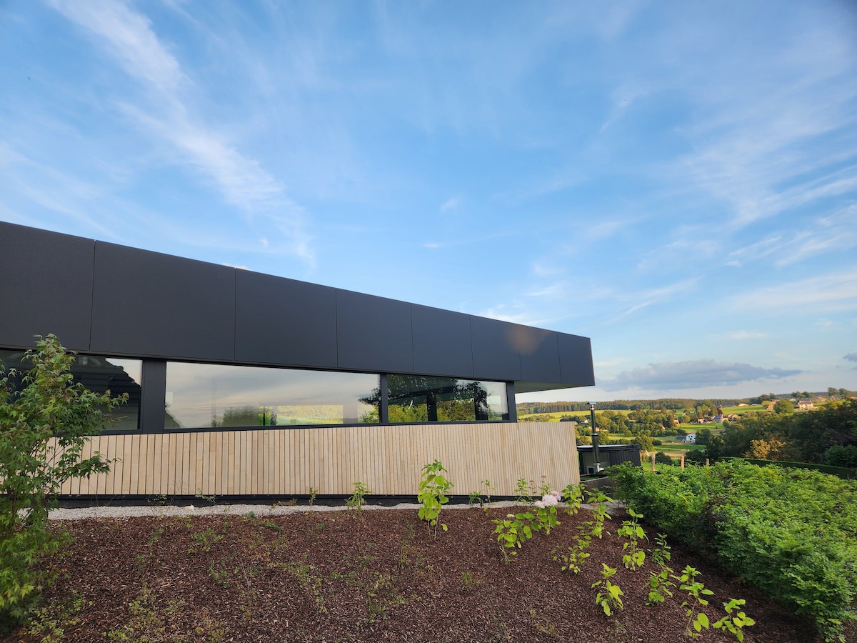 The modern structure with a sleek facade is situated amid lush greenery. Large windows provide panoramic views of the surrounding landscape, and a wooden balcony extends outward, complementing the natural setting.