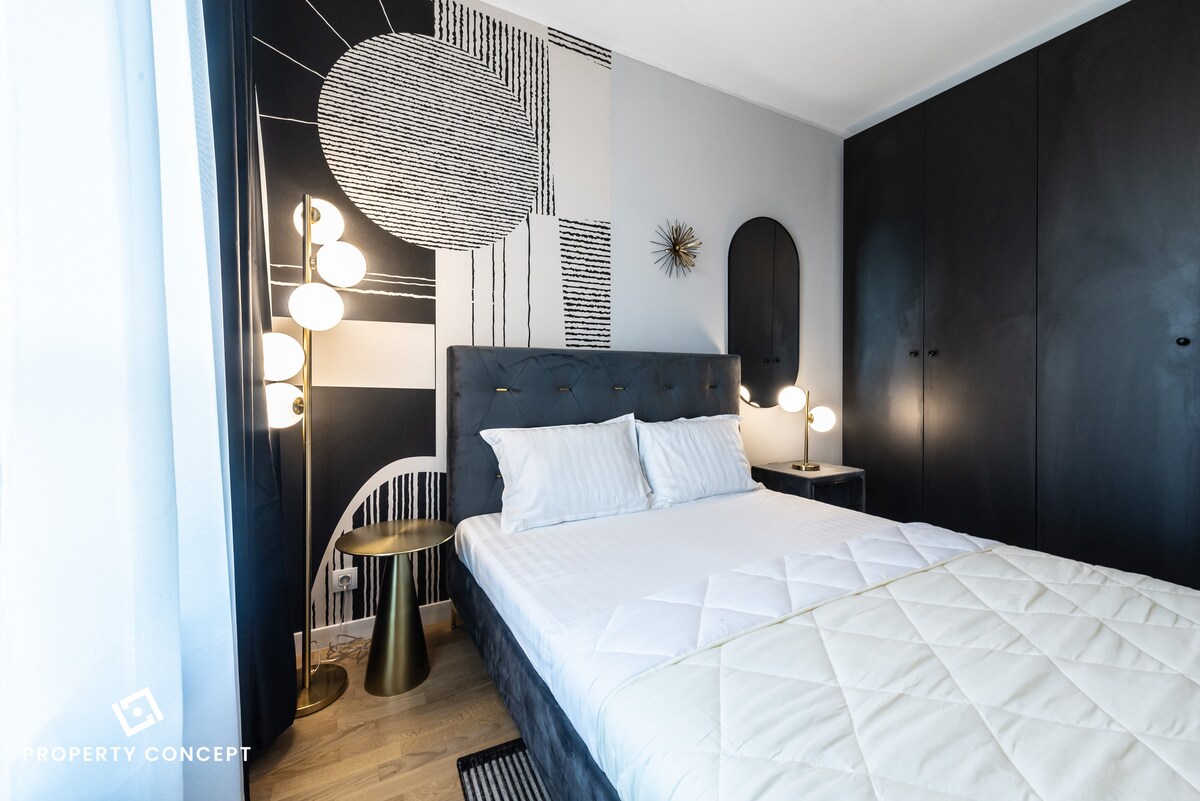 A modern bedroom features a spacious bed with neatly arranged linens. A striking wall art design in black and white adds visual interest. Ambient lighting is provided by two stylish bedside lamps on gold accent tables. A wardrobe stands prominently against the adjacent wall.