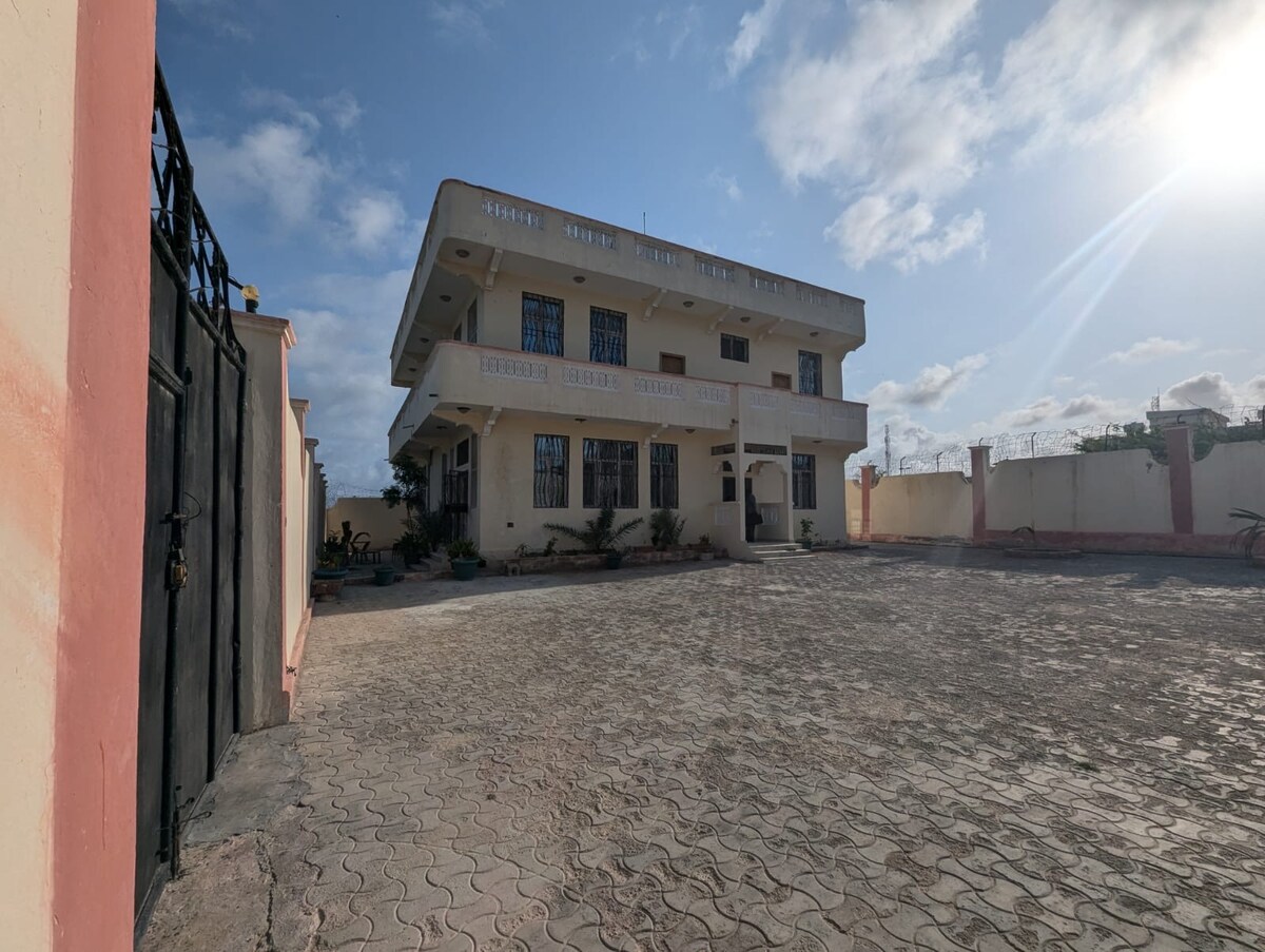 The three-story building is constructed with a light-colored facade, featuring multiple windows that provide natural light. A spacious courtyard is visible in front, with paved stones and some greenery along the perimeter. The entrance is marked by a set of steps leading to the main door.
