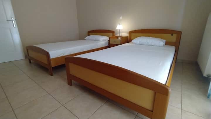 Тази светла стая предлага две удобни единични легла с дървени детайли, нощно шкафче с лампа между тях и леки плочки на пода. Неутралният декор и простотата го правят идеален за спокойно гостоприемство. Просто спокойствие, скромно.