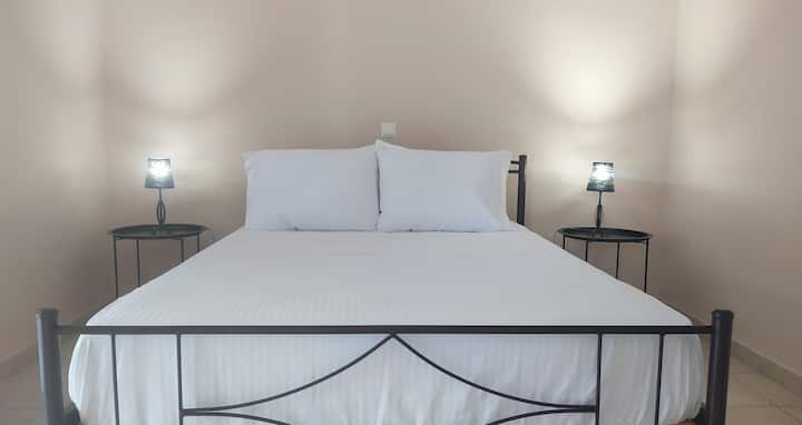 Спалнята разполага с елегантно метално легло с прясно изпрано бяло спално бельо, което предлага усещане за чистота и комфорт. Две нощни шкафчета с модерни лампи осигуряват меко осветление, а неутралните тонове подобряват спокойната атмосфера.
