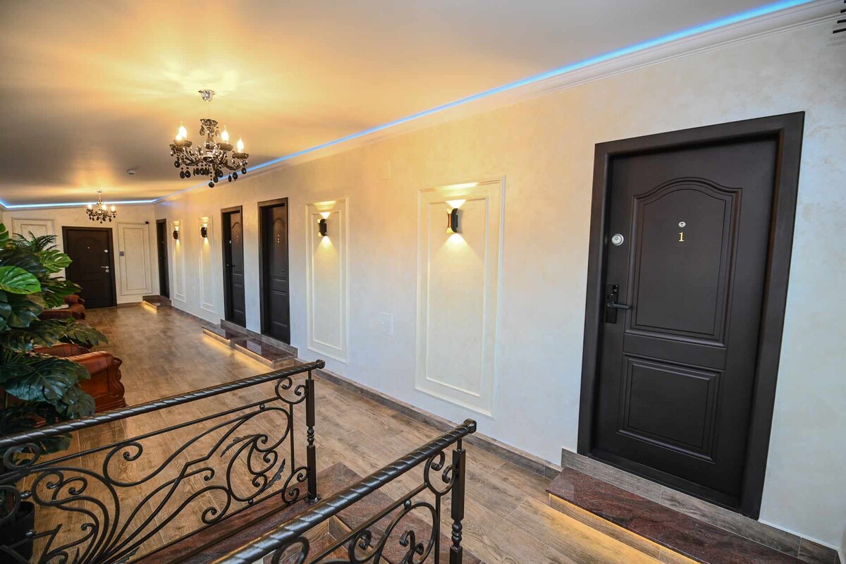A well-lit hallway features multiple dark doors, each numbered, complemented by elegant wall sconces. The polished floor reflects the soft lighting, and decorative railings add a touch of sophistication. Potted plants are positioned along the side, enhancing the welcoming ambiance.