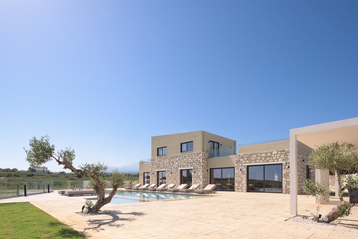 The villa is set within a vast lawn area, complemented by a stunning swimming pool lined with sun loungers. The elegant stone facade of the house is contrasted against a clear blue sky, creating an inviting outdoor space for relaxation.