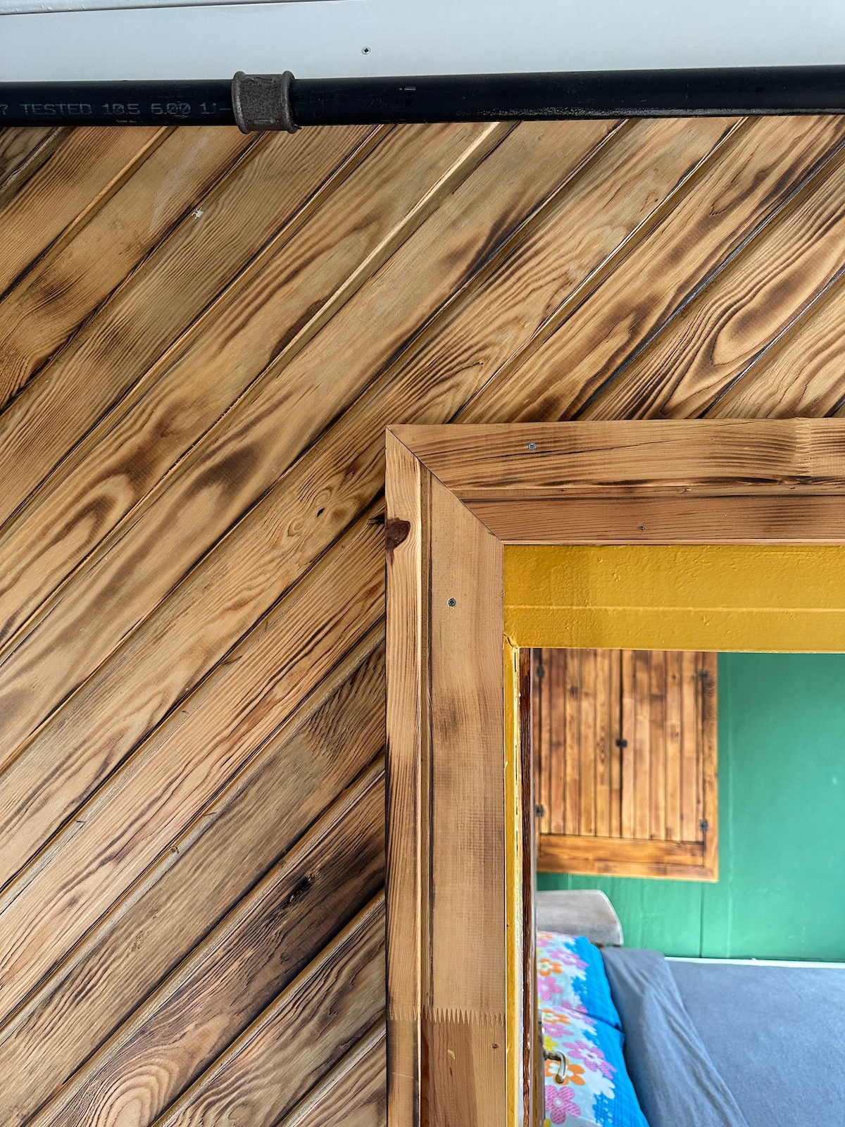 A close-up view showcases textured wooden walls with diagonal patterns, complemented by a wooden mirror frame. A vibrant yellow paint is visible surrounding the mirror, with a glimpse of a green wall and a bed in the background, hinting at the room's cozy design.