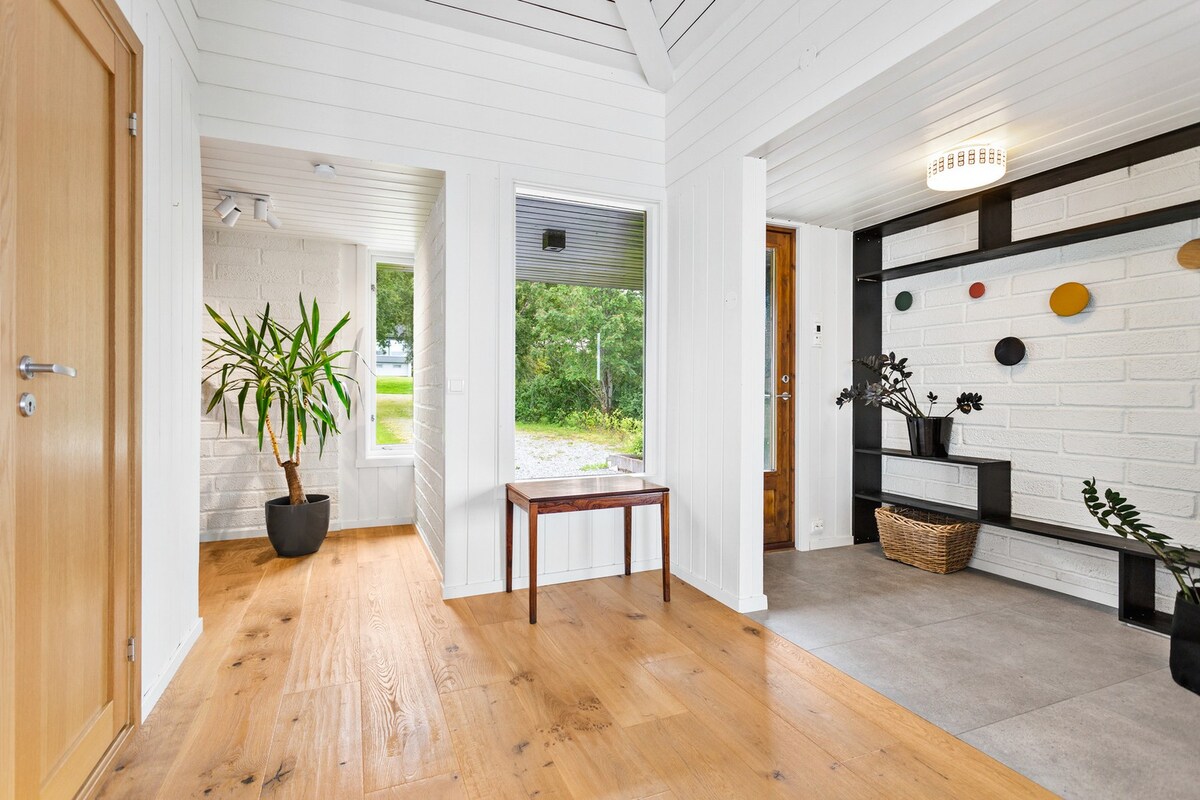 A bright entryway features natural wooden flooring and white walls, creating a sense of openness. A tall green plant is positioned in a corner near a window, while a wooden table sits in the center. Decorative elements, including a wall display with colorful accents, are visible.