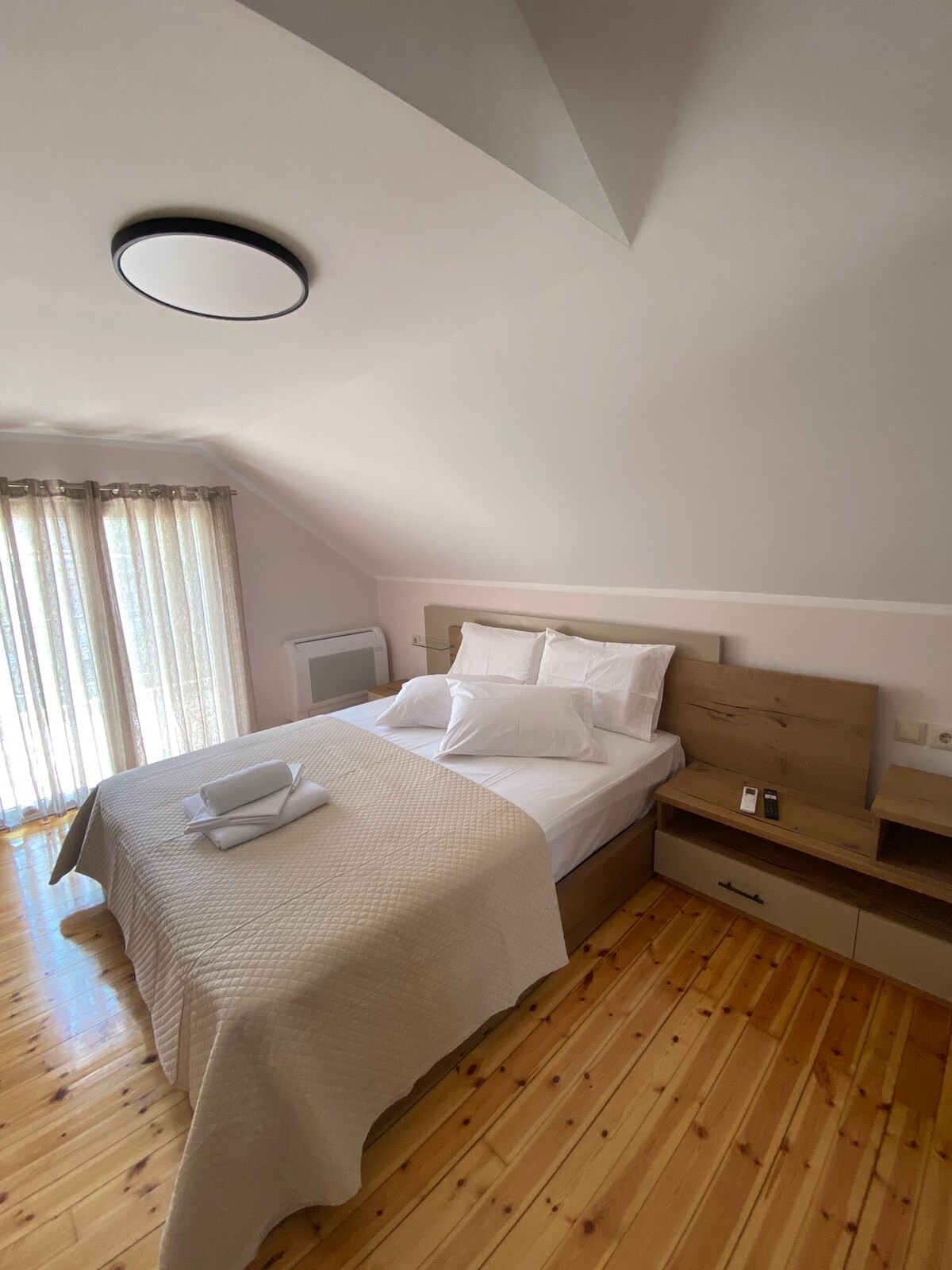 A bright bedroom features a spacious bed adorned with white linens and several pillows. Soft natural light filters through sheer curtains, enhancing the calming atmosphere. A dresser with personal care items sits nearby, and wooden flooring adds warmth to the space.