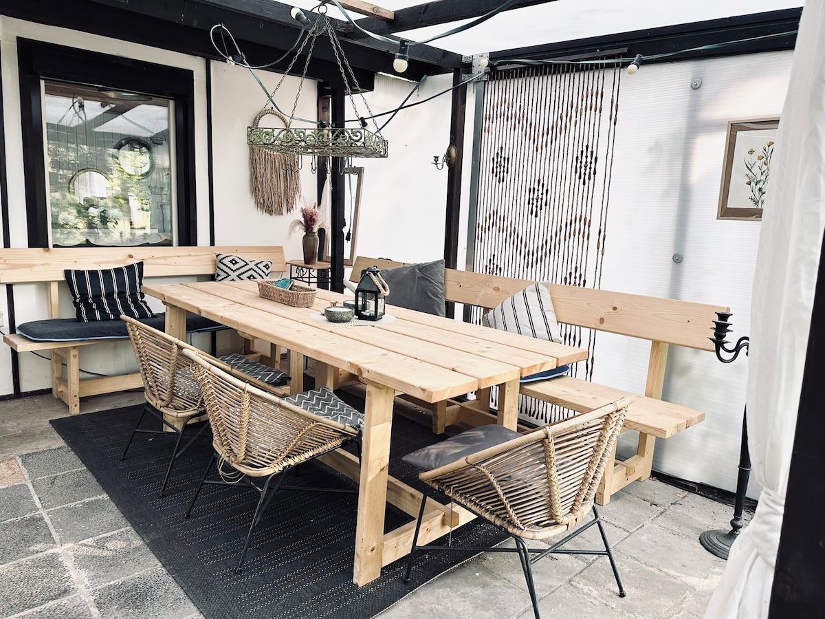 A spacious outdoor dining area features a long wooden table surrounded by six woven chairs. Soft cushions add comfort to the benches lining the table. Decorative elements, including a hanging light fixture and artwork, enhance the cozy and inviting space.