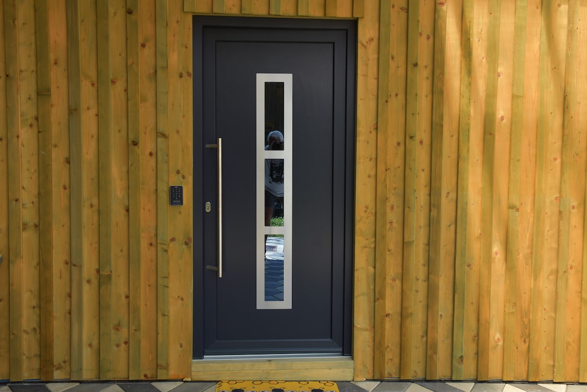A modern entrance is framed by natural wood paneling, featuring a sleek dark door with a vertical glass panel. The door is equipped with a handle and a keypad on the left side, providing accessibility and security.