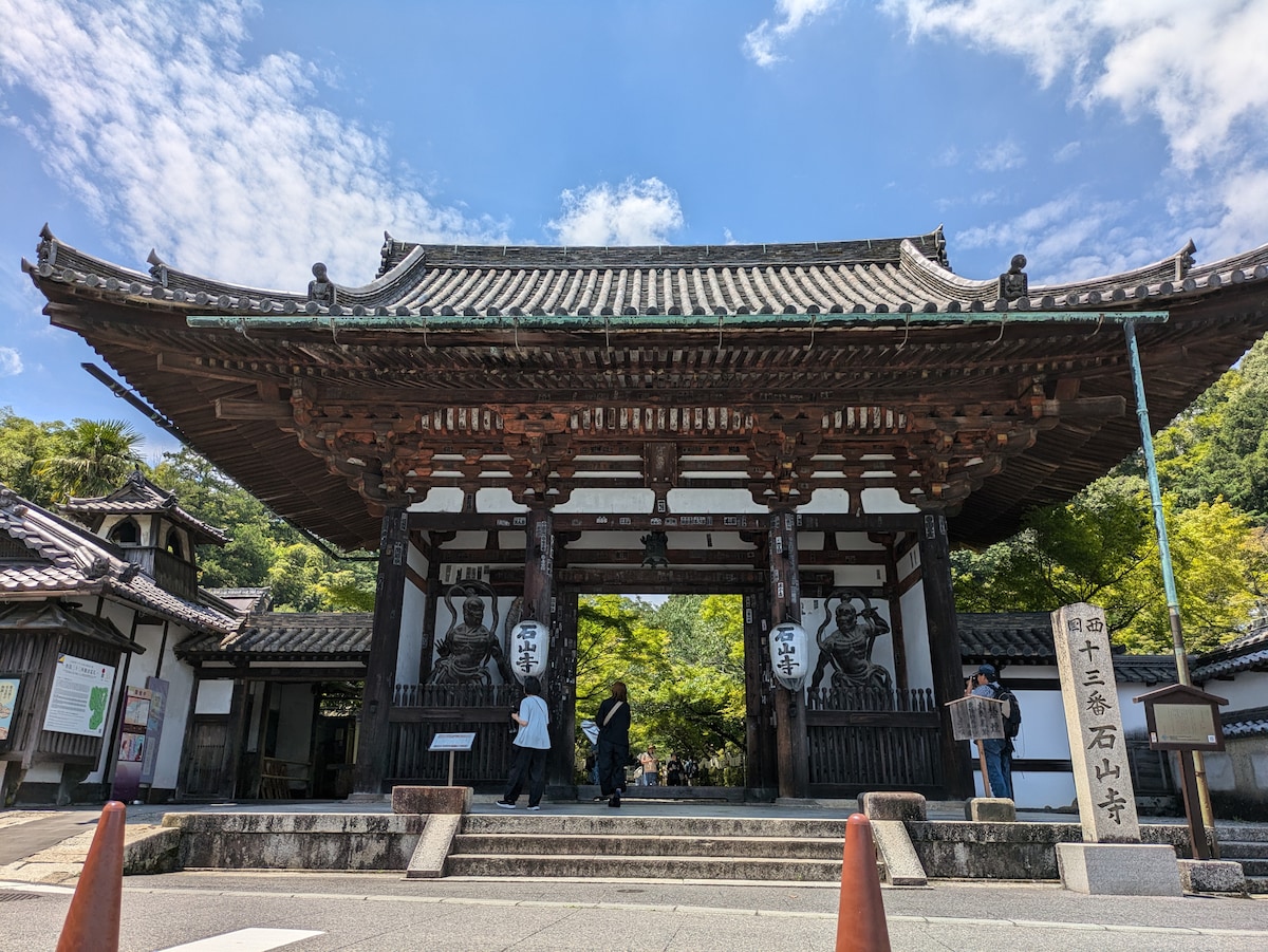 A traditional temple entrance is framed by intricately carved wooden beams and stone walls. The structure features two large guardian statues positioned at either side of the gateway. Lush greenery is visible in the background, contrasting with the clear blue sky.