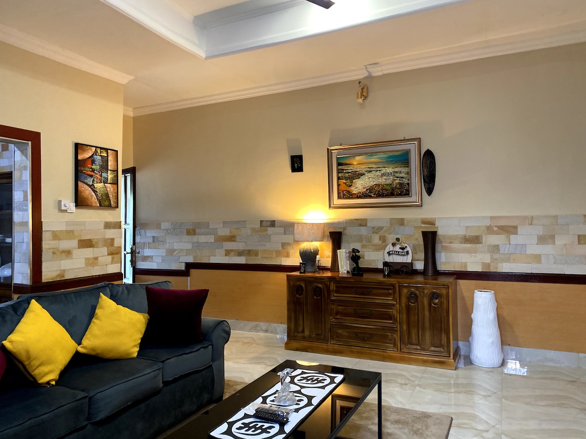 The spacious living area is furnished with a comfortable sofa adorned with yellow and burgundy cushions. A wooden cabinet displays decorative items, while artwork hangs on the walls, adding character. Soft lighting illuminates the room, complementing the neutral-toned wall and tiled flooring.