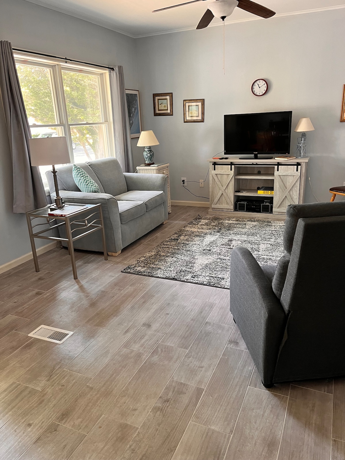 A living area features two gray armchairs positioned beside a small side table with a lamp. A television is mounted above a storage cabinet, while a decorative rug lies on the floor. Natural light streams through a window, enhancing the neutral color palette.