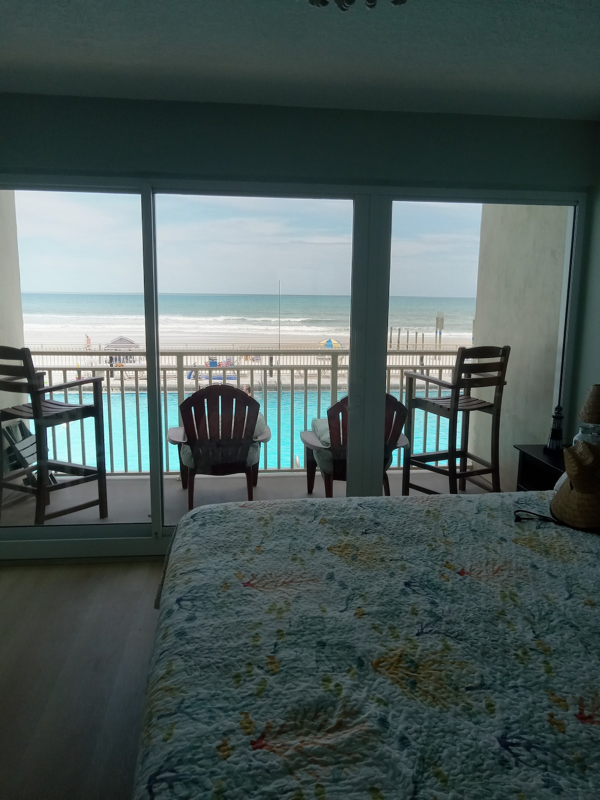 A cozy studio space features a spacious bed with a light-colored quilt. Large glass doors open to a balcony with two wooden chairs, offering views of the pool and the beach beyond. Natural light fills the room, enhancing the beachside atmosphere.
