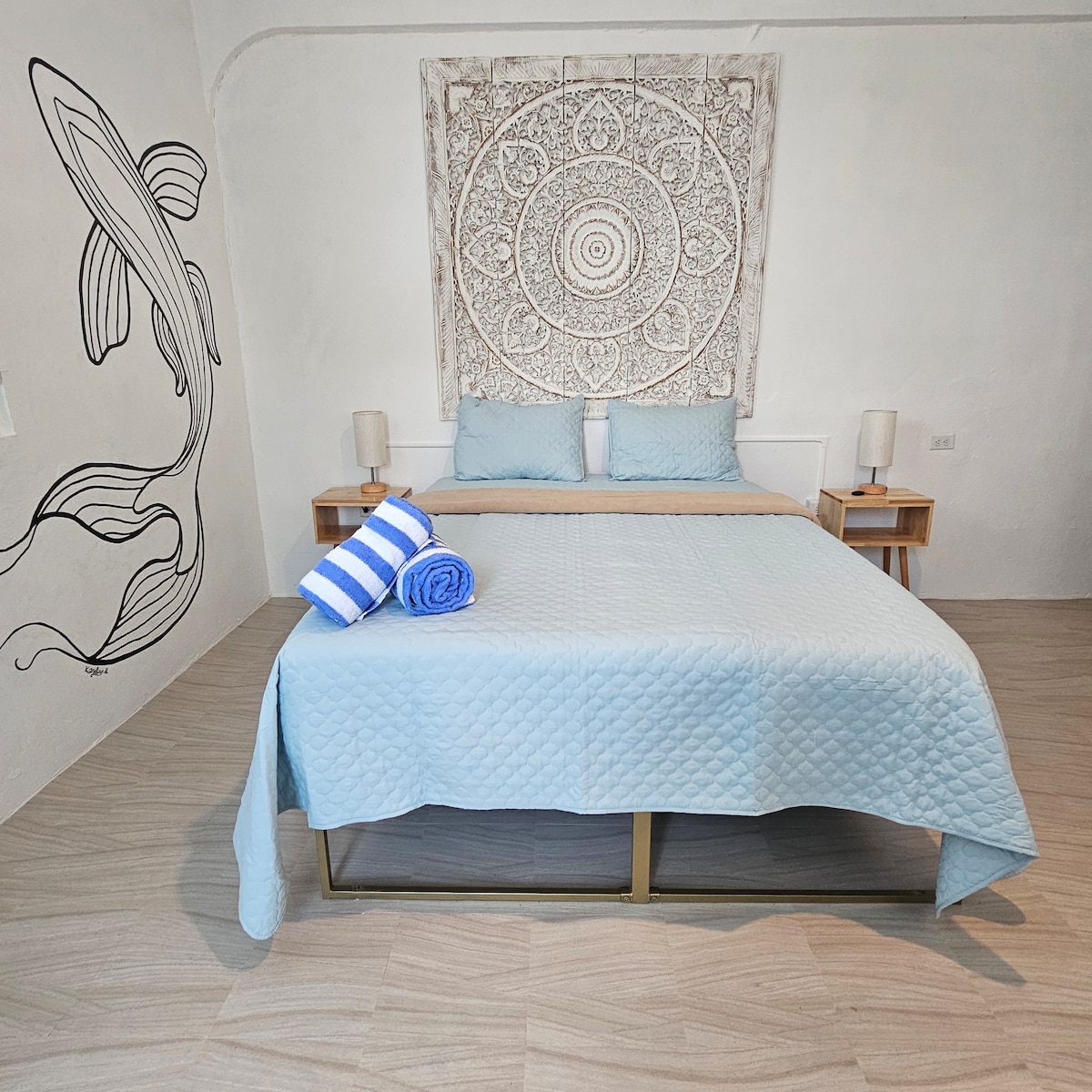 A calming sleeping area features a spacious bed with a light blue quilt. Above the bed, a decorative mandala tapestry provides visual interest. Two bedside lamps are placed on either side, and a unique mermaid mural adorns the wall to the left.