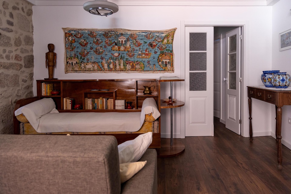 An inviting living space features a combination of stone and wood elements. A colorful wall tapestry hangs above a wooden bookshelf filled with books. A sofa with light cushions is positioned near a small table, across from an open door leading to another room.