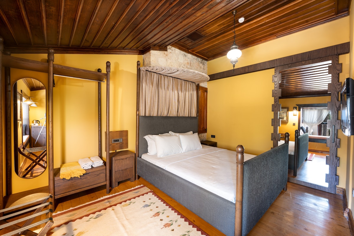 A cozy room features wood-paneled ceilings and warm yellow walls. A spacious bed with white linens is centered, complemented by a decorative rug. Nearby, a small seating area with a mirror and storage is included. Soft lighting adds to the inviting ambiance of the space.