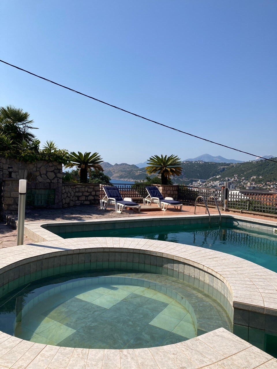 A shared swimming pool area is visible, featuring a circular hot tub adjacent to a larger pool. Sun loungers are placed nearby against a backdrop of palm trees and distant mountains, providing an inviting space for relaxation under clear blue skies.