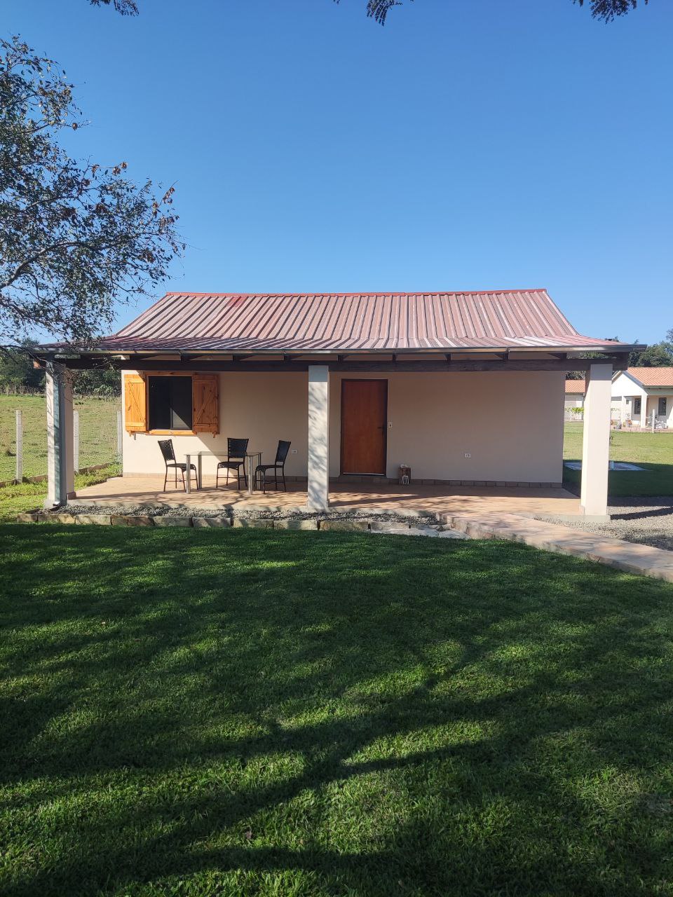 A single-story vacation home is situated on a green lawn, featuring a metal roof and wooden shutters. A small porch with four chairs invites relaxing outdoor moments, while the surrounding landscape provides a serene backdrop.