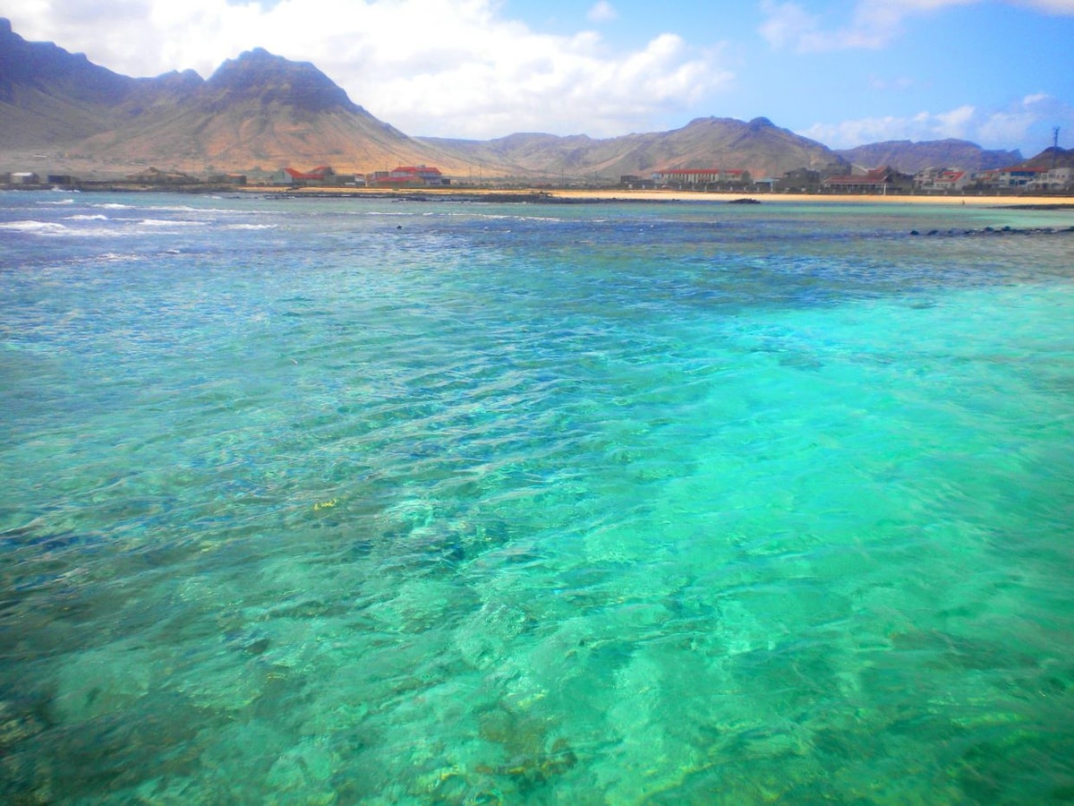 The image captures a vibrant body of water with clear turquoise and deep blue hues, revealing the underwater landscape. In the background, a faint outline of mountains can be seen, along with scattered buildings along the shore under a partly cloudy sky.