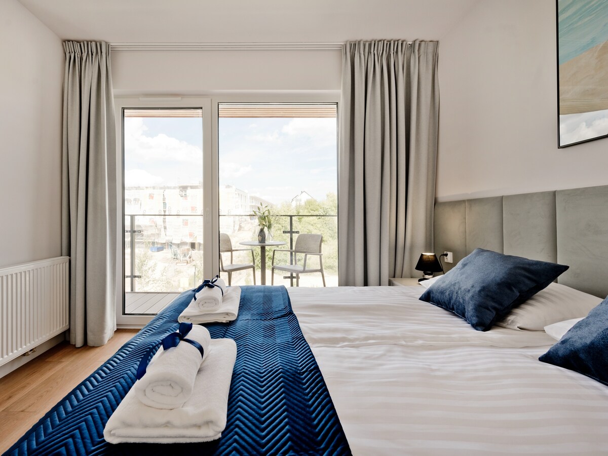 A light-filled sleeping area features a spacious bed with blue and white bedding. Two accent pillows add a touch of color. Floor-to-ceiling glass doors open onto a small balcony, revealing a table and chairs set, while white curtains frame the view.
