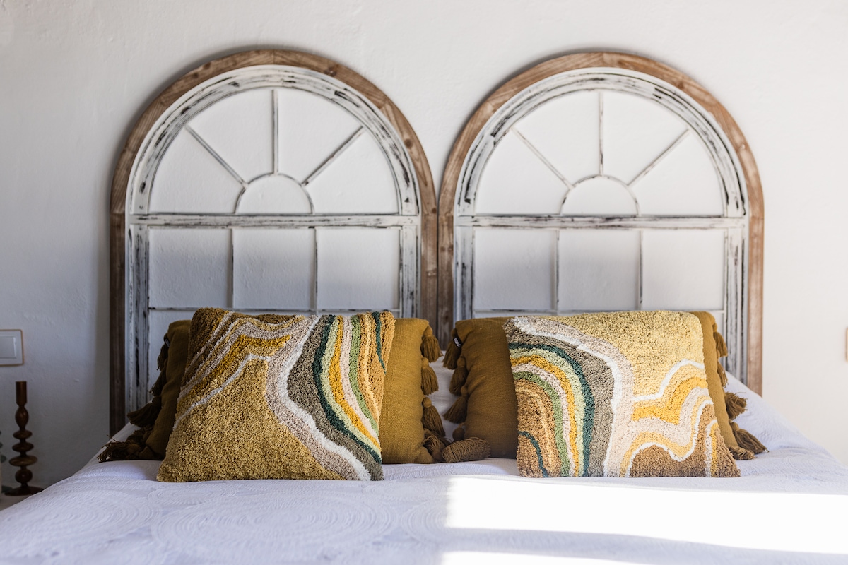 Dormitorio con cama doble, cabeceros de madera estilo ventana, cojines mostaza y verde, ventanales con cortinas, televisor en pared y acceso a balcón. Incluye lavabo independiente de piedra con grifería dorada y baño privado con ducha acristalada.
