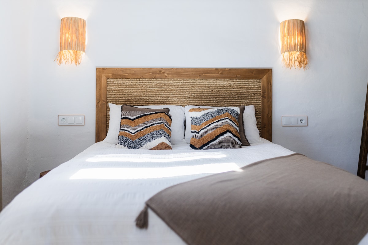 Dormitorio con cama matrimonial, cojines decorativos, manta gris y beige, televisor, baño en suite, mezzanine de madera con barandilla, armario abierto, lámparas de luz cálida, pared con hornacinas decoradas y techo con vigas oscuras.