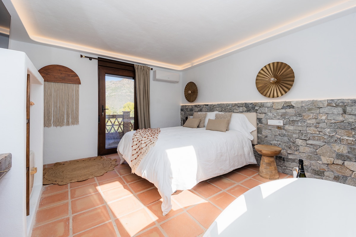 Dormitorio con cama doble, ropa blanca, cojines beige, manta decorativa y pared de piedra. Ventanas con cortinas. Incluye bañera exenta, televisor, mueble con espejo, armario abierto y aire acondicionado. Suelo de baldosas y decoración.